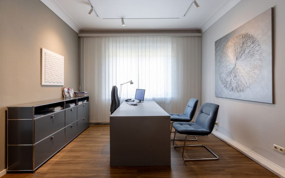 Raum einer Arztpraxis. Mittig steht ein schwerer Tisch. Rechts zwei Stühle, links einer. Links ist ein Sideboard. Minrath USM Akustikdecke Fenix