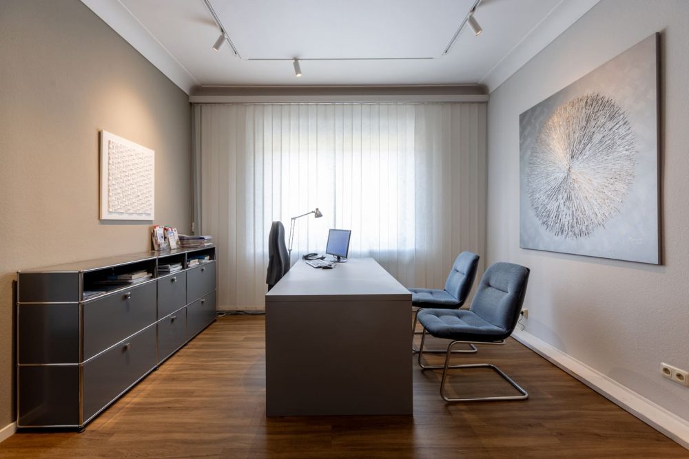 Raum einer Arztpraxis. Mittig steht ein schwerer Tisch. Rechts zwei Stühle, links einer. Links ist ein Sideboard. Minrath USM Akustikdecke Fenix