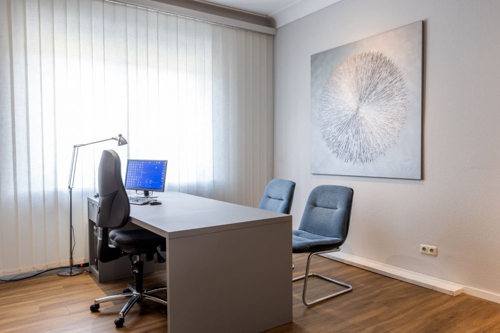 Ansicht eines Arztbesprechungszimmers mit einem Schreibtisch in der Mitte mit einem Bildschirm darauf. Links des Tisches steht ein Schreibtischstuhl. Rechts davon zwei Stühle. Minrath Fenix