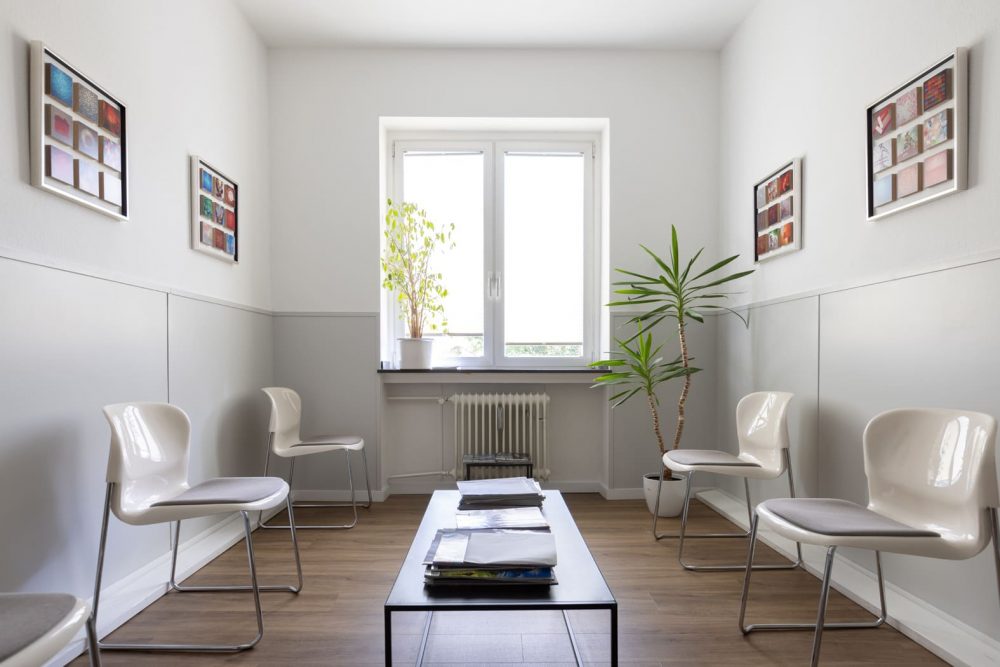Wartebereich einer Arztpraxis. Mittig steht ein Tisch mit Zeitschriften darauf. Hinten im Bild ist ein Fenster mit einer Topfpflanze auf dem Fensterbrett. Im rechten Eck steht eine Yukkapalme. Links und rechts im Raum stehen Stühle. Minrath Wandvertäfelung